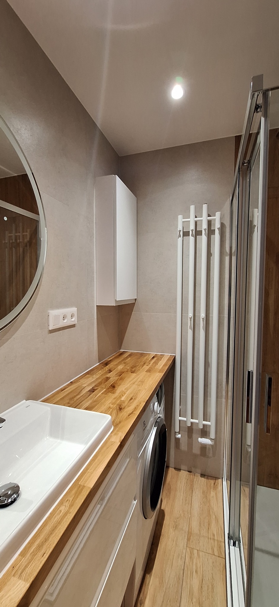 Nowoczesna łazienka z drewnianym blatem, białą umywalką nablatową, pralką pod blatem, grzejnikiem drabinkowym i kabiną prysznicową z przezroczystego szkła.