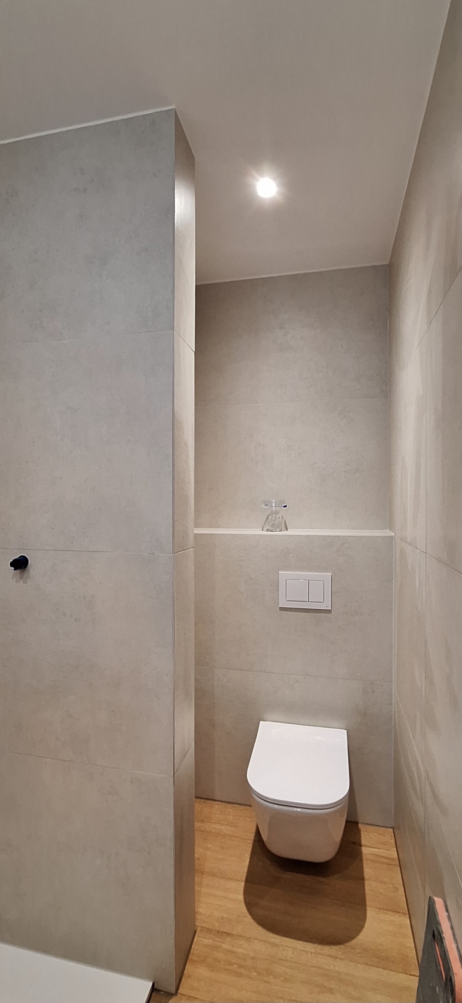 Nowoczesna, minimalistyczna łazienka z podwieszaną toaletą, płytkami w odcieniach szarości i drewnopodobną podłogą, akcent dekoracyjny stanowi szklana ozdoba.