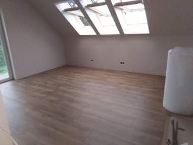 Poddasze z nową, jasną podłogą panelową, okna dachowe w skosie, białe ściany z widocznymi punktami elektrycznymi, grzejnik owinięty folią ochronną w rogu.