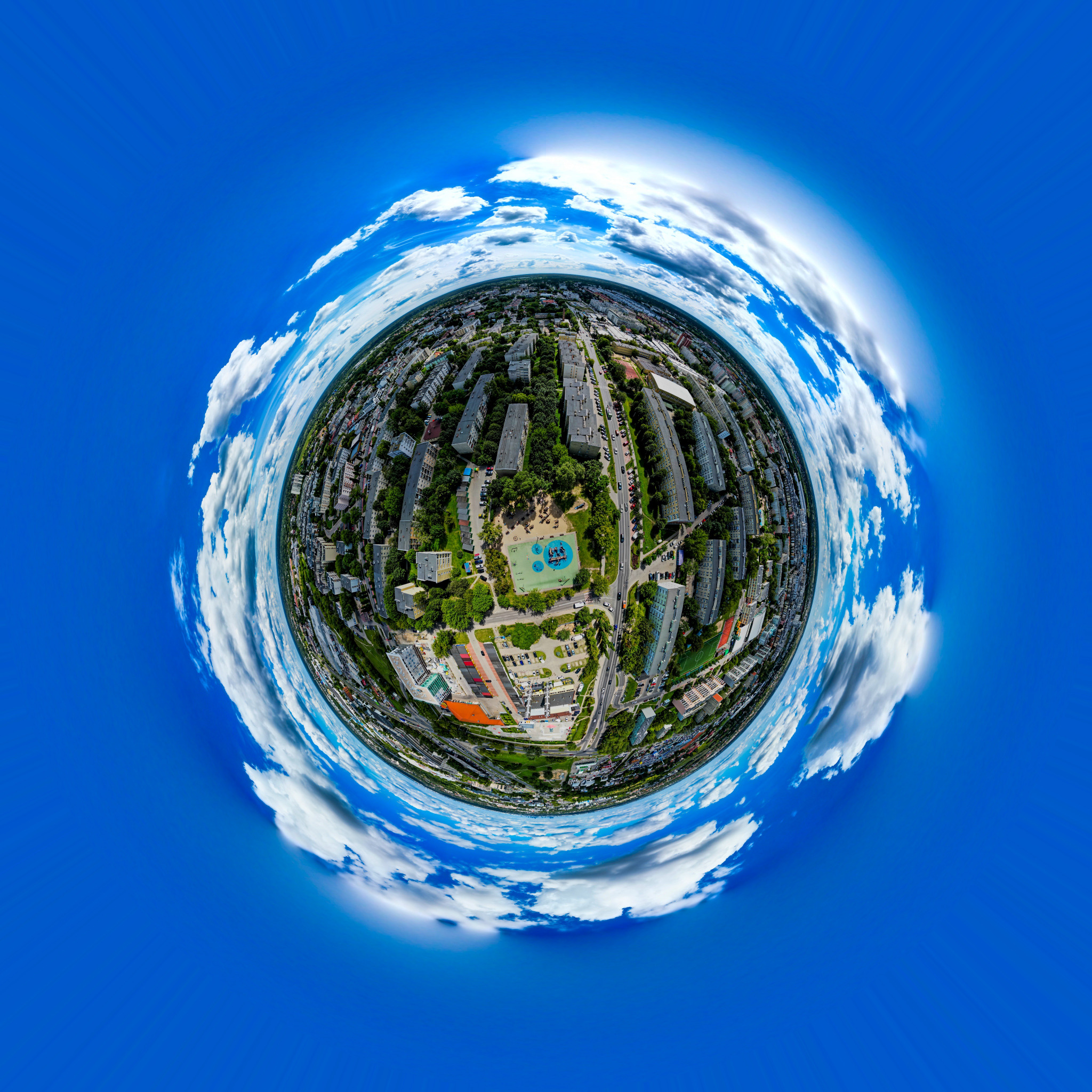 Panoramiczne ujęcie sferyczne miasta z widokiem na budynki mieszkalne, park z placem zabaw i ulice, otoczone błękitnym niebem z białymi chmurami.
