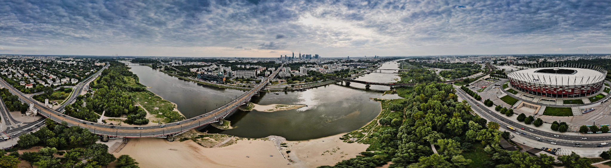 Panoramiczny widok z lotu ptaka na Warszawę, Wisłę z mostami, Stadion Narodowy i panoramę miasta pod pochmurnym niebem.
