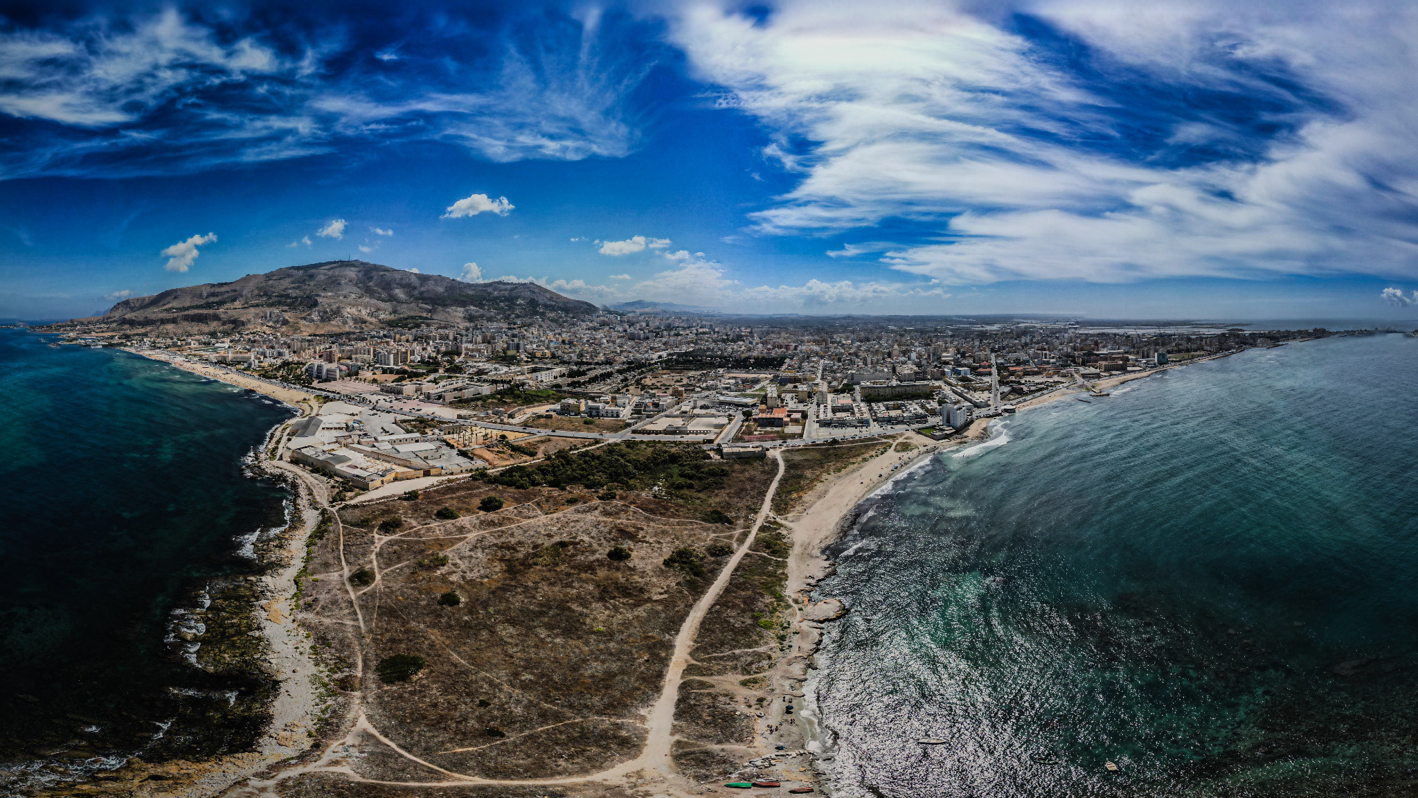 Panoramiczny widok z lotu ptaka na miasto położone nad brzegiem morza, z wyraźnie zaznaczonymi budynkami, plażą i górującą górą w tle pod błękitnym niebem z chmurami.