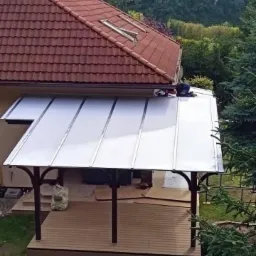 Pracownik montuje zadaszenie z poliwęglanu nad tarasem, widoczny fragment dachu domu z dachówką ceramiczną i oknami dachowymi.