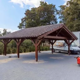 Drewniana wiata garażowa z brązową dach&oacute;wką ceramiczną na posesji z kostki brukowej, na tle zieleni i zaparkowanych samochod&oacute;w.