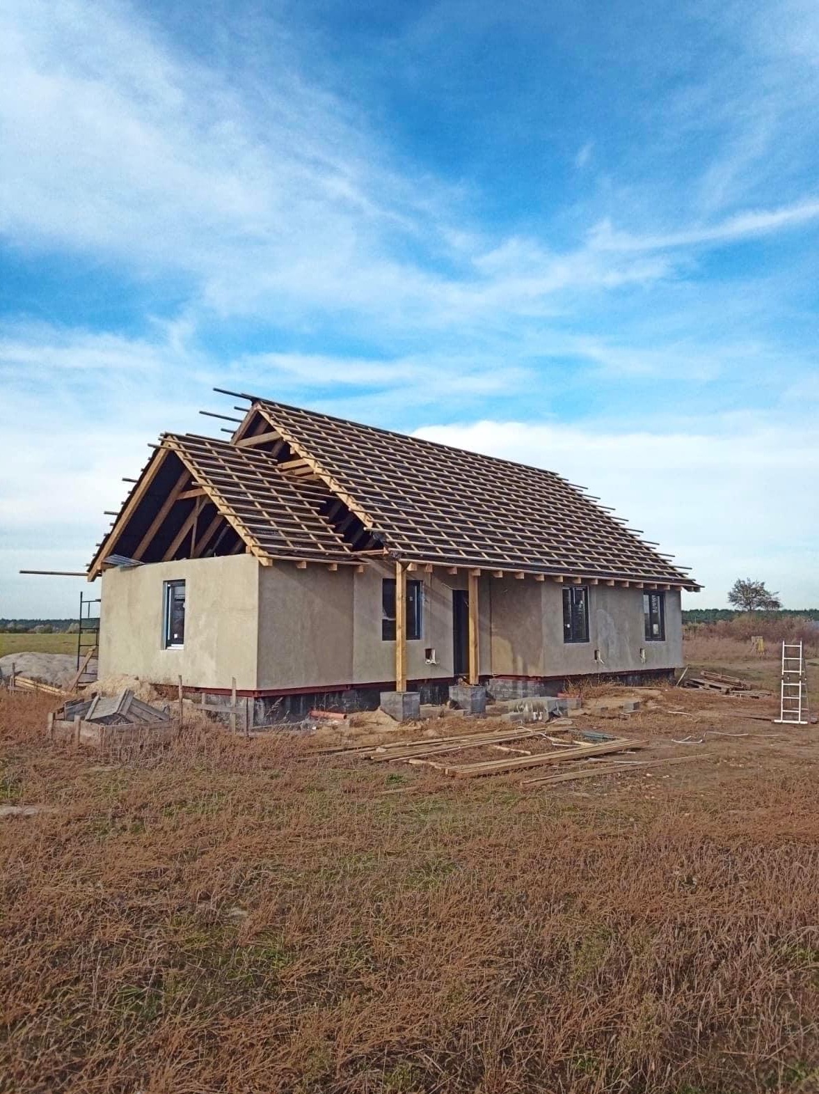 Surowy dom jednorodzinny w trakcie budowy, z widoczną więźbą dachową, oknami w czarnych ramach i szarym tynkiem zewnętrznym, otoczony polami i błękitnym niebem.