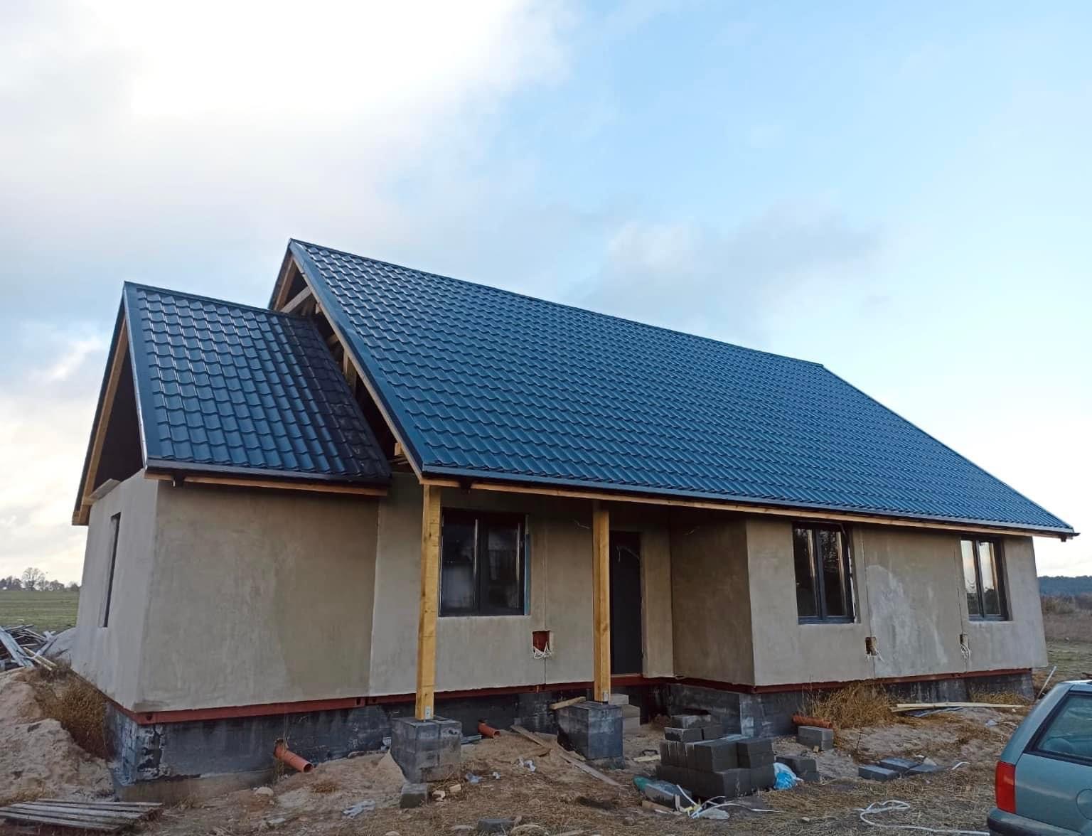 Dom w trakcie budowy z nowym, ciemnoniebieskim dachem z blachodachówki, widoczne drewniane słupy podtrzymujące okap i surowy tynk na ścianach.