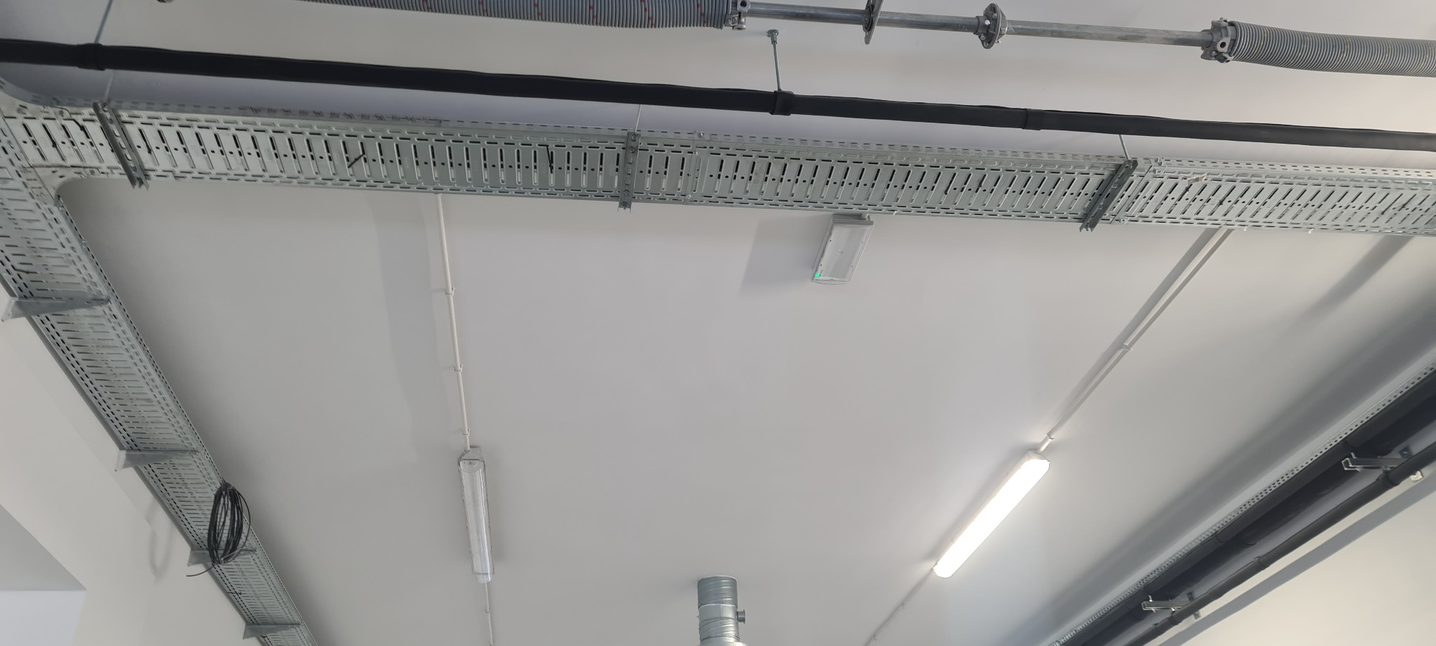 Widok z dołu na biały sufit z zamontowanymi korytkami kablowymi, rurami i oświetleniem fluorescencyjnym w hali przemysłowej.