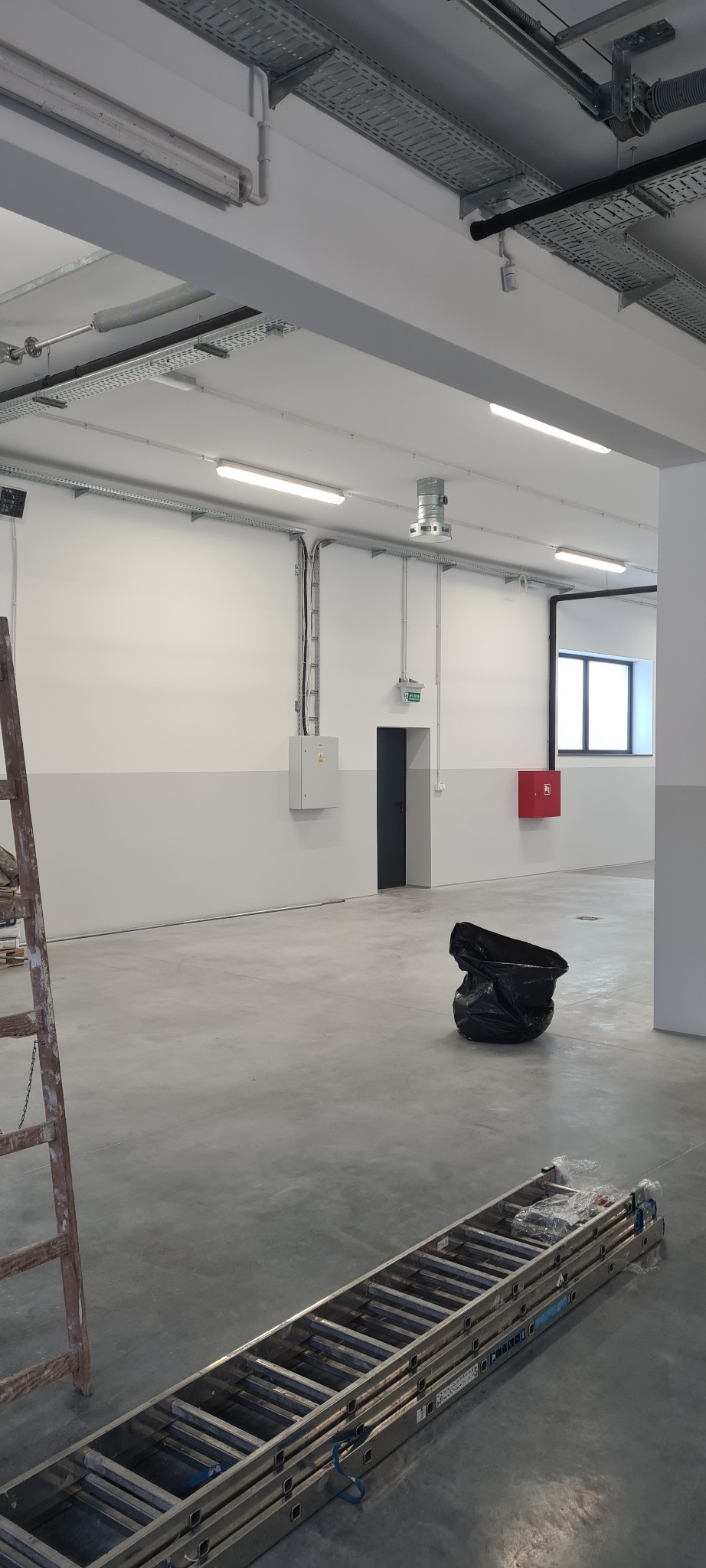 Wnętrze hali produkcyjnej z widocznymi instalacjami elektrycznymi pod sufitem, skrzynką elektryczną na ścianie, drabinami i workiem na śmieci na podłodze.