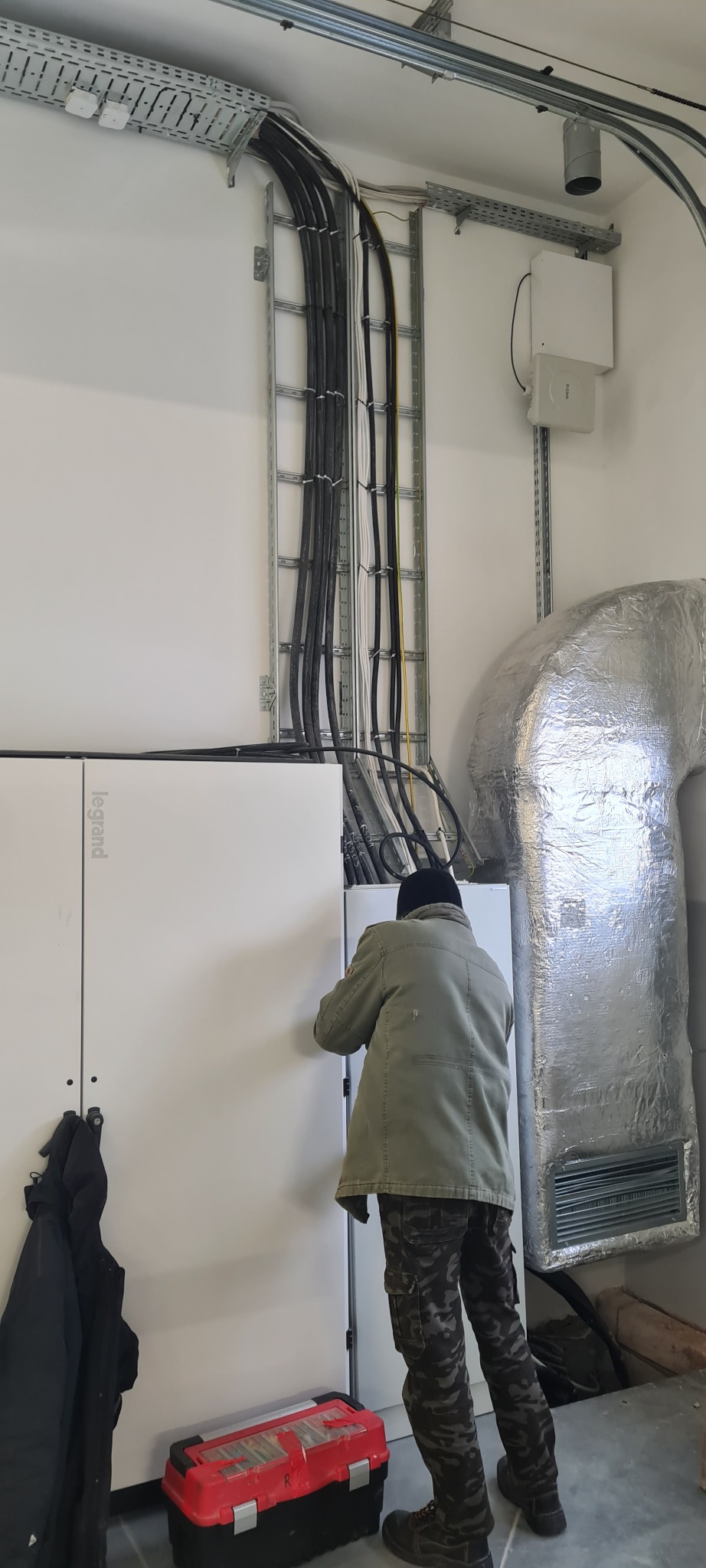 Elektryk w zielonej kurtce i moro spodniach montuje białą skrzynkę elektryczną Legrand, widoczne kable w korytkach, czerwona skrzynka z narzędziami i srebrny kanał wentylacyjny.