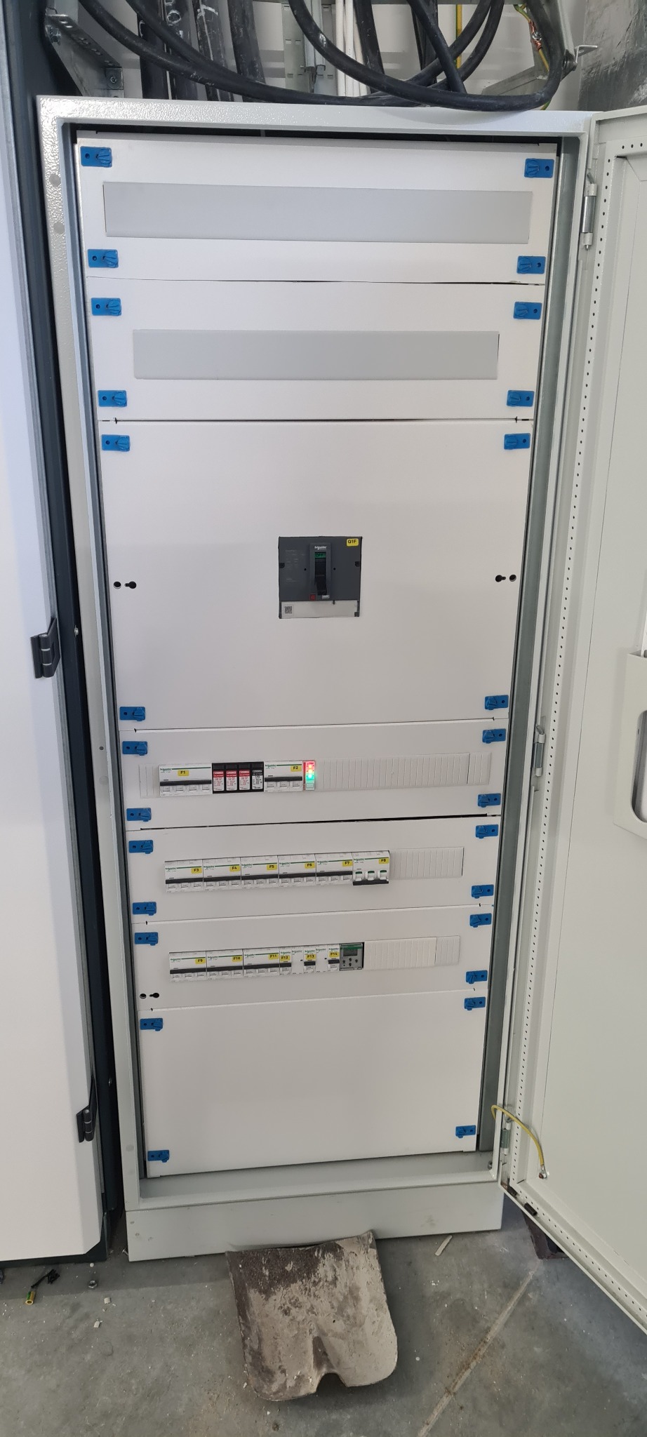 Otwarta szafa elektryczna z widocznymi zabezpieczeniami, wyłącznikiem głównym Schneider Electric i listwami zaciskowymi; widoczne kable w górnej części szafy; na podłodze płyta betonowa