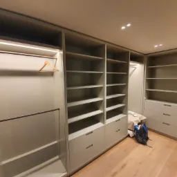 Wnętrze garderoby z szarymi szafami wnękowymi z półkami i szufladami, widoczny wieszak na ubrania, na podłodze leży plecak.