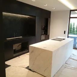 Wnętrze kuchni w trakcie montażu z wyspą wykończoną białym marmurem, czarne szafki kuchenne z podświetleniem LED, widoczne zabezpieczenia podłogi.