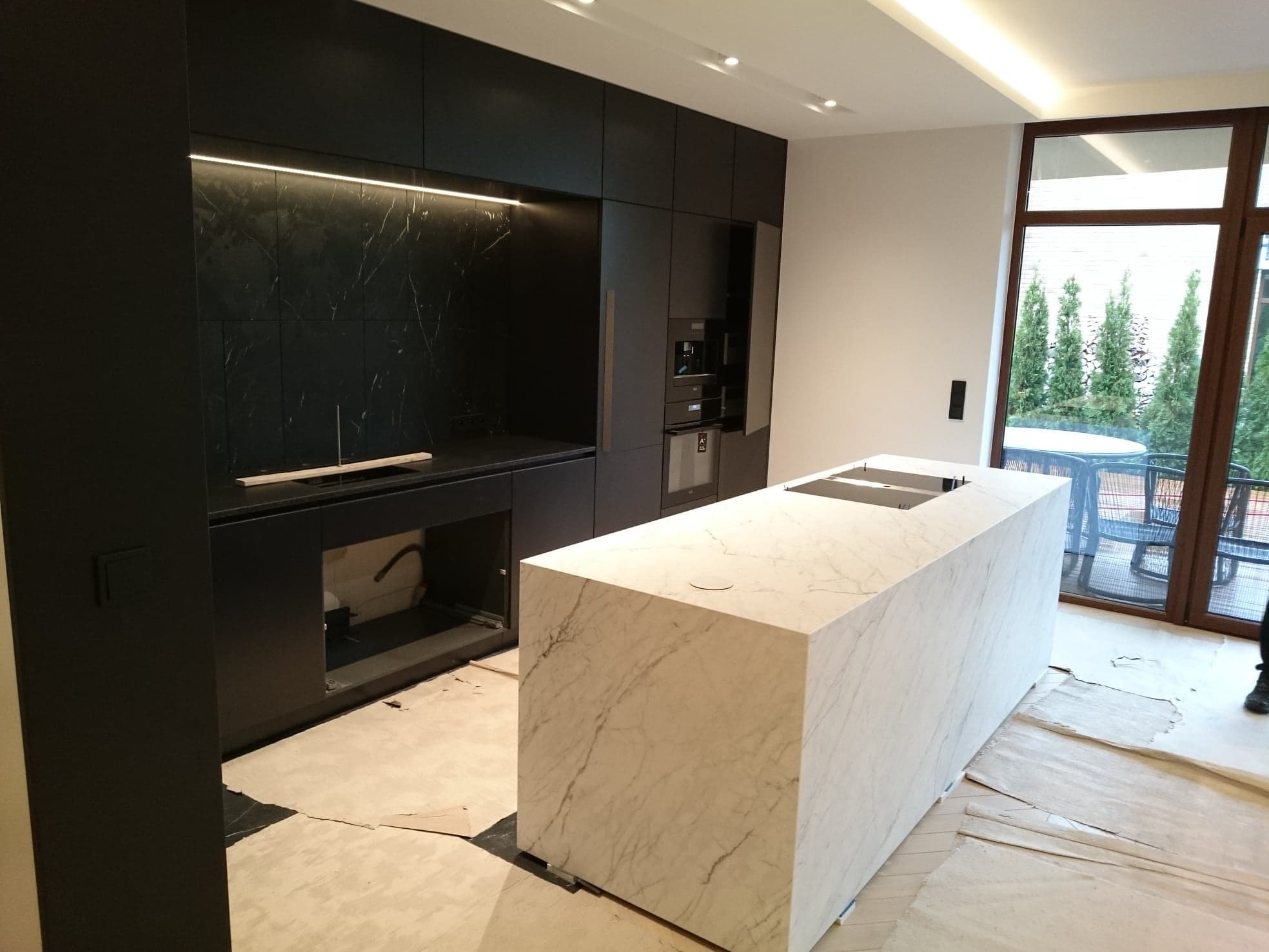 Wnętrze kuchni w trakcie montażu z wyspą wykończoną białym marmurem, czarne szafki kuchenne z podświetleniem LED, widoczne zabezpieczenia podłogi.