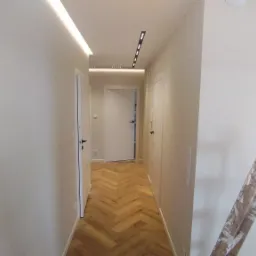 Korytarz w trakcie prac, widoczna drabina malarska, ściany przygotowane do malowania, parkiet ułożony w jodełkę, oświetlenie sufitowe.