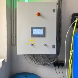 Panel sterowania Siemens Simatic HMI z zieloną lampką sygnalizacyjną, przycisk awaryjny, okablowanie i niebieski zbiornik na wodę w tle.