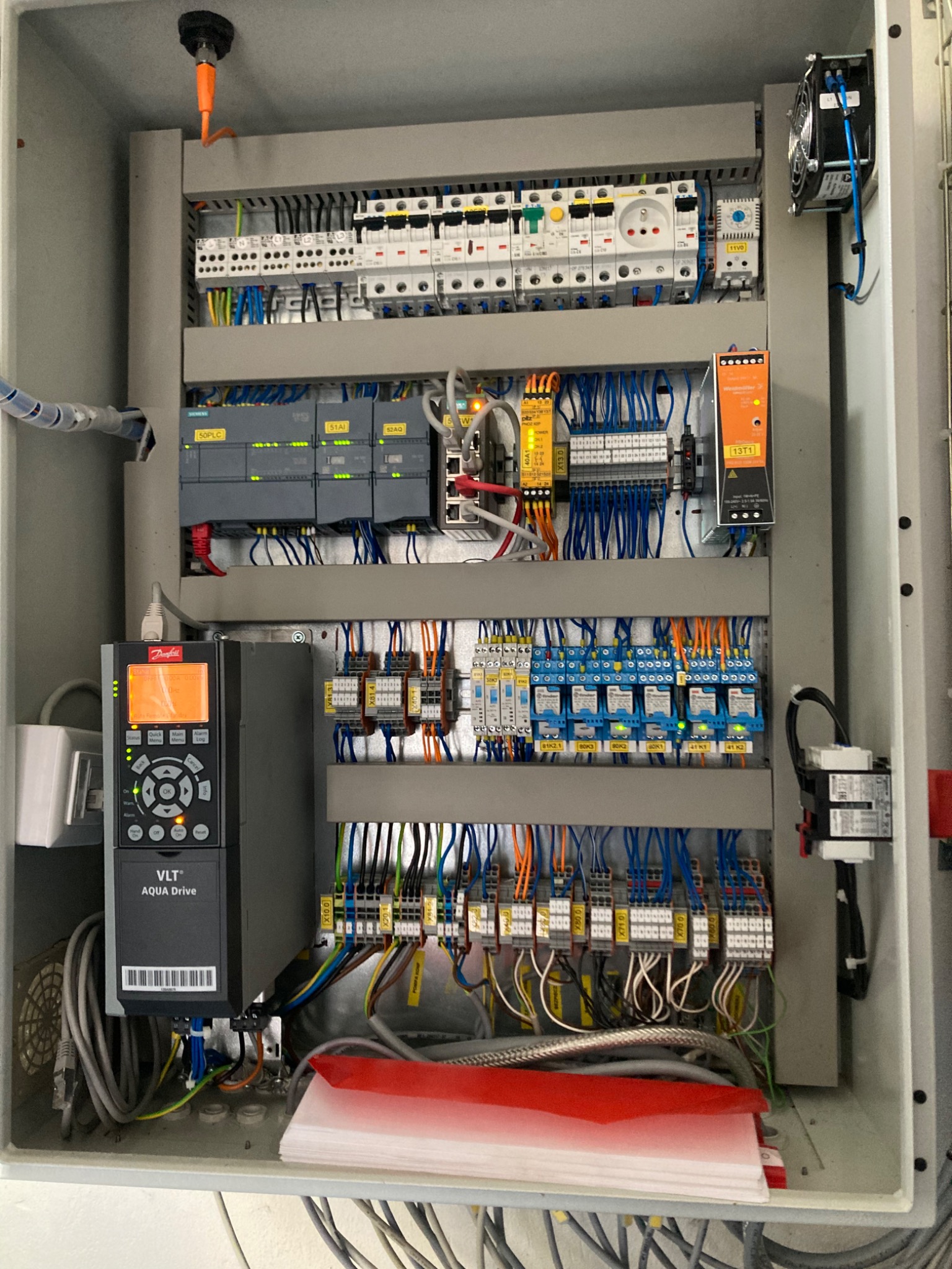 Otwarta szafa sterownicza z modułami Siemens, Danfoss VLT Aqua Drive, bezpiecznikami i licznymi przewodami w kolorach pomarańczowym i niebieskim, wentylator oraz dokumentacja techniczna w dolnej...