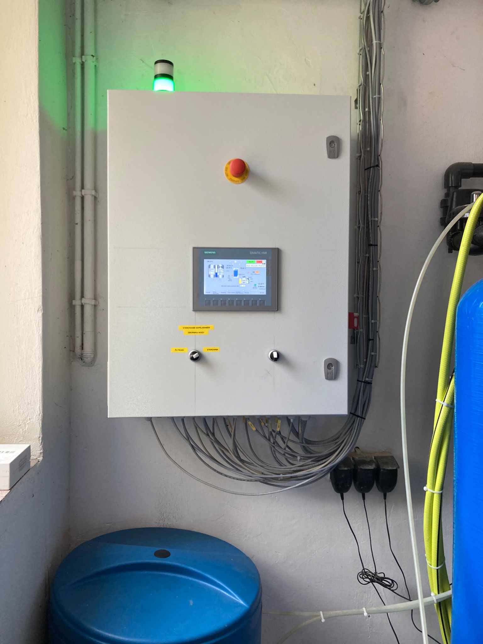 Panel sterowania Siemens Simatic HMI z zieloną lampką sygnalizacyjną, przycisk awaryjny, okablowanie i niebieski zbiornik na wodę w tle.