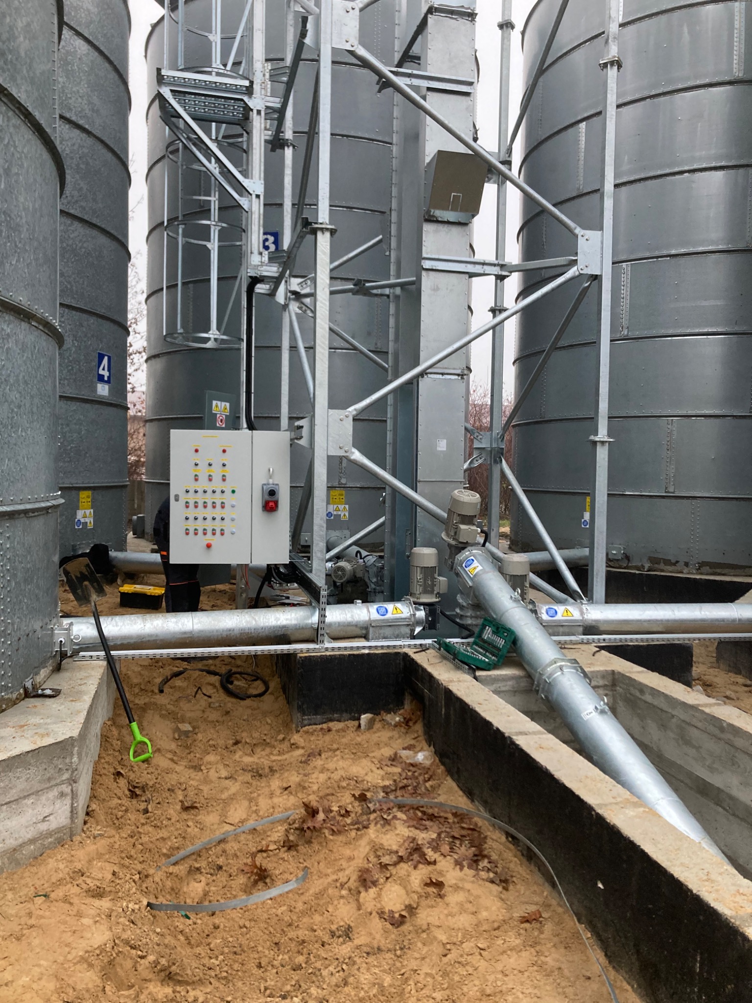 Instalacja elektryczna i rurociągowa przy silosach zbożowych, widoczna szafa sterownicza, rury, silniki, narzędzia i fragment pracownika w kombinezonie.