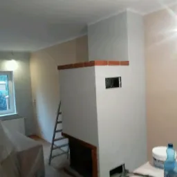 Wnętrze po remoncie z widocznym kominem z ceglanym wykończeniem, drabina oparta o ścianę, widoczny fragment okna i zakryte meble.