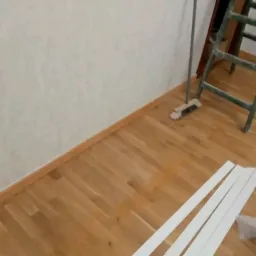 Przygotowane do montażu białe listwy przypodłogowe leżą na parkiecie, w tle drabina malarska oparta o ścianę z delikatnym wzorem.