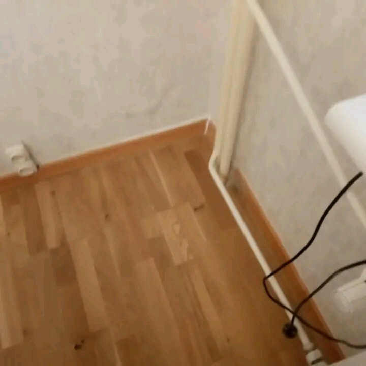 Fragment łazienki z drewnianą podłogą, widoczne rury i przyłącza, ściany z jasną tapetą.