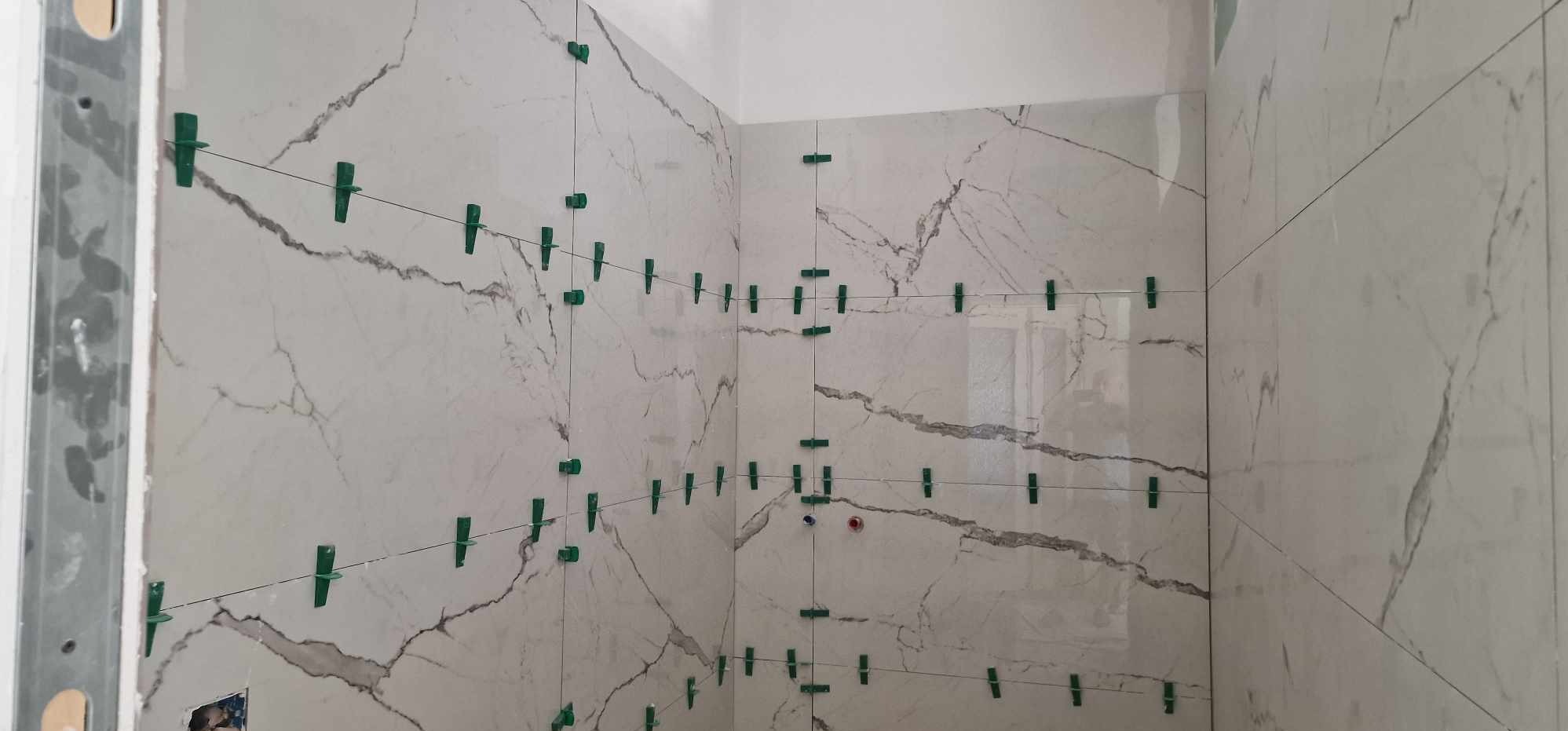 Ściana w trakcie układania płytek imitujących marmur, z użyciem zielonych klipsów poziomujących i sznurka do wyznaczania linii. Widoczne fragmenty metalowej konstrukcji ściany.