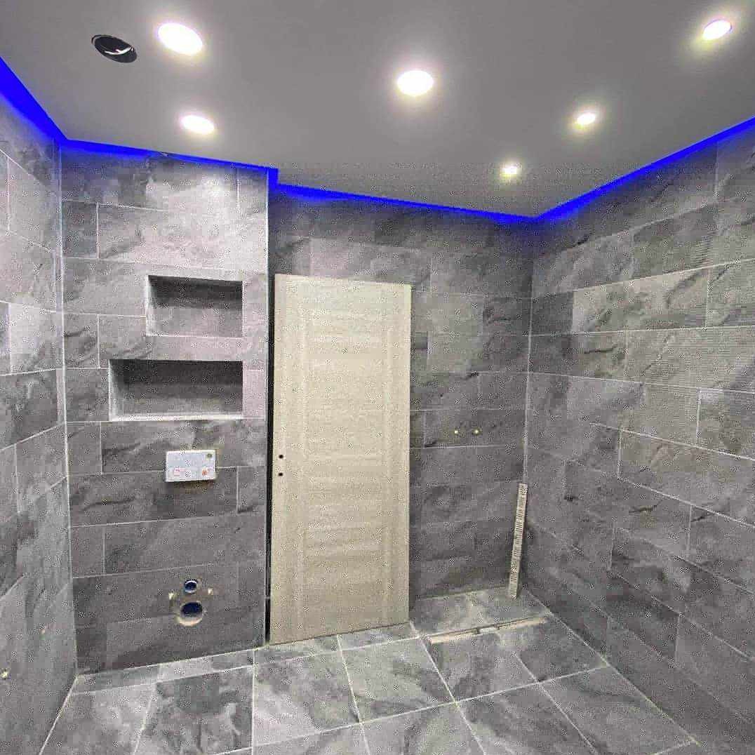 Szara łazienka z podświetleniem LED na suficie, widoczne wnęki w ścianie wyłożonej płytkami, białe drzwi i częściowo ułożona podłoga z płytek imitujących kamień.