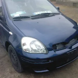 Niebieski samochód osobowy z uszkodzonym przodem, widoczny brakujący grill i uszkodzona tablica rejestracyjna na tle ziemistej nawierzchni.