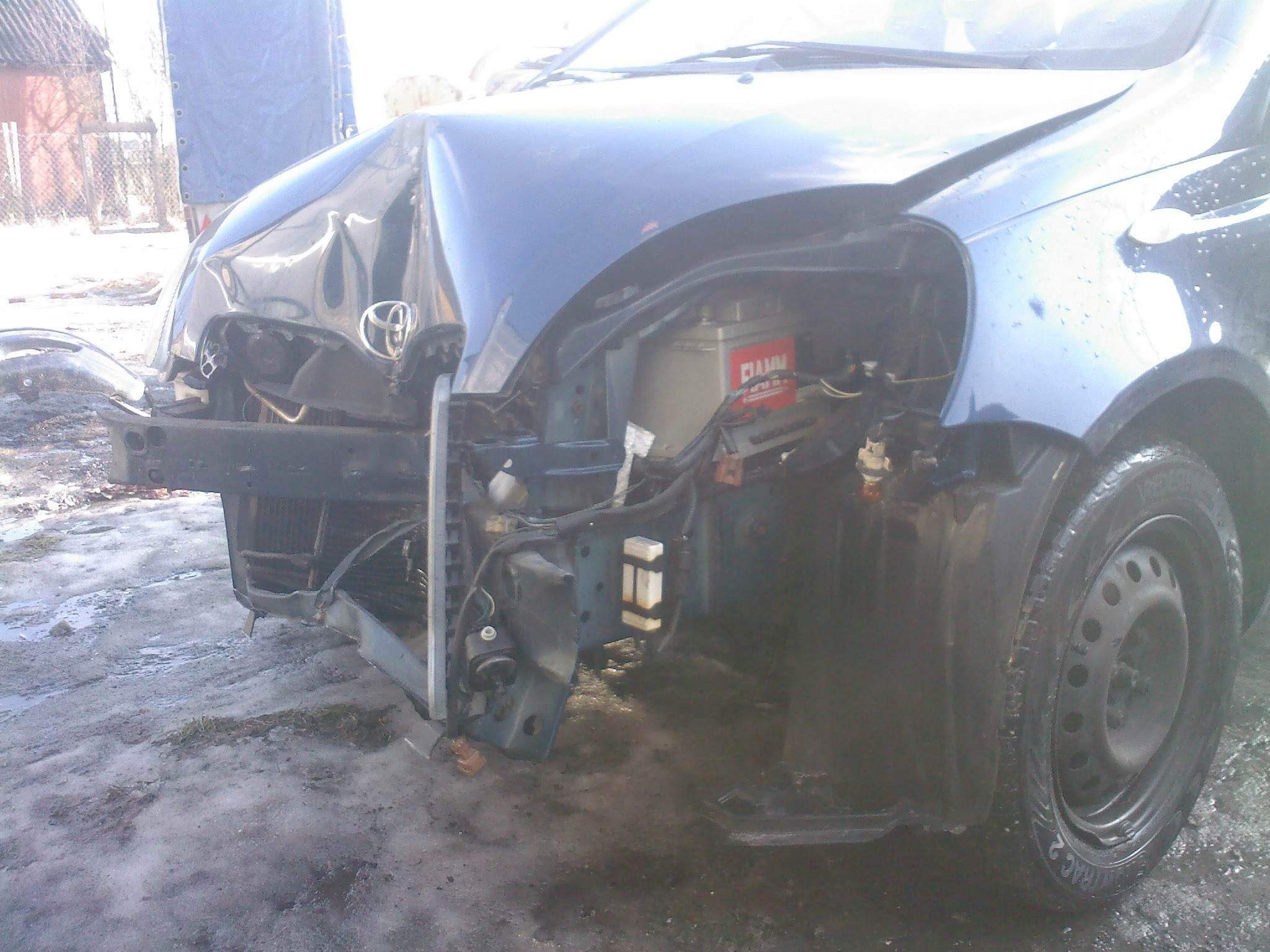 Uszkodzony przód niebieskiego samochodu Toyota Yaris po kolizji, widoczna zgnieciona maska, brakujący reflektor i odsłonięty akumulator, na ziemi resztki błota i lodu.