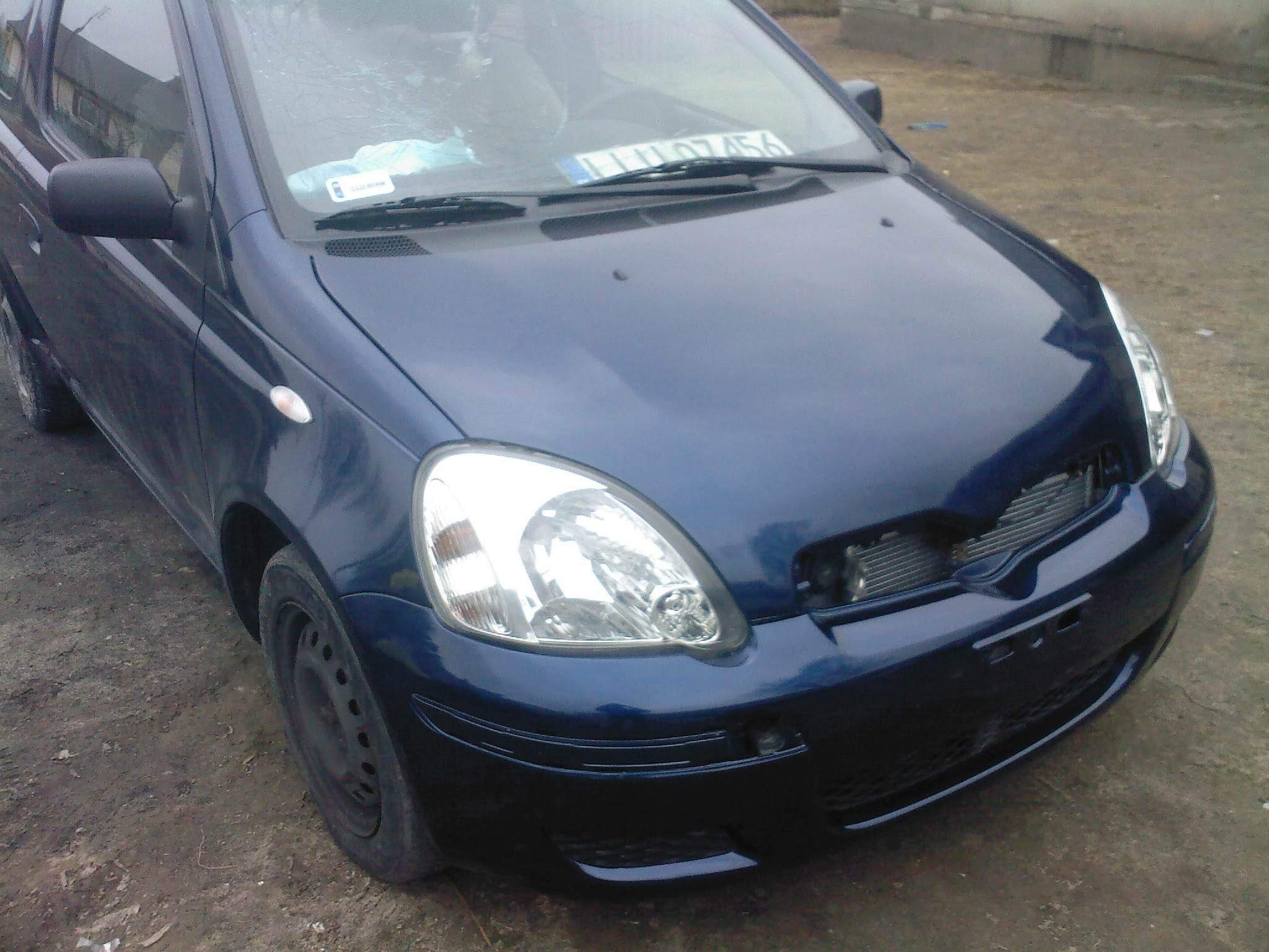Niebieski samochód osobowy z uszkodzonym przodem, widoczny brakujący grill i uszkodzona tablica rejestracyjna na tle ziemistej nawierzchni.