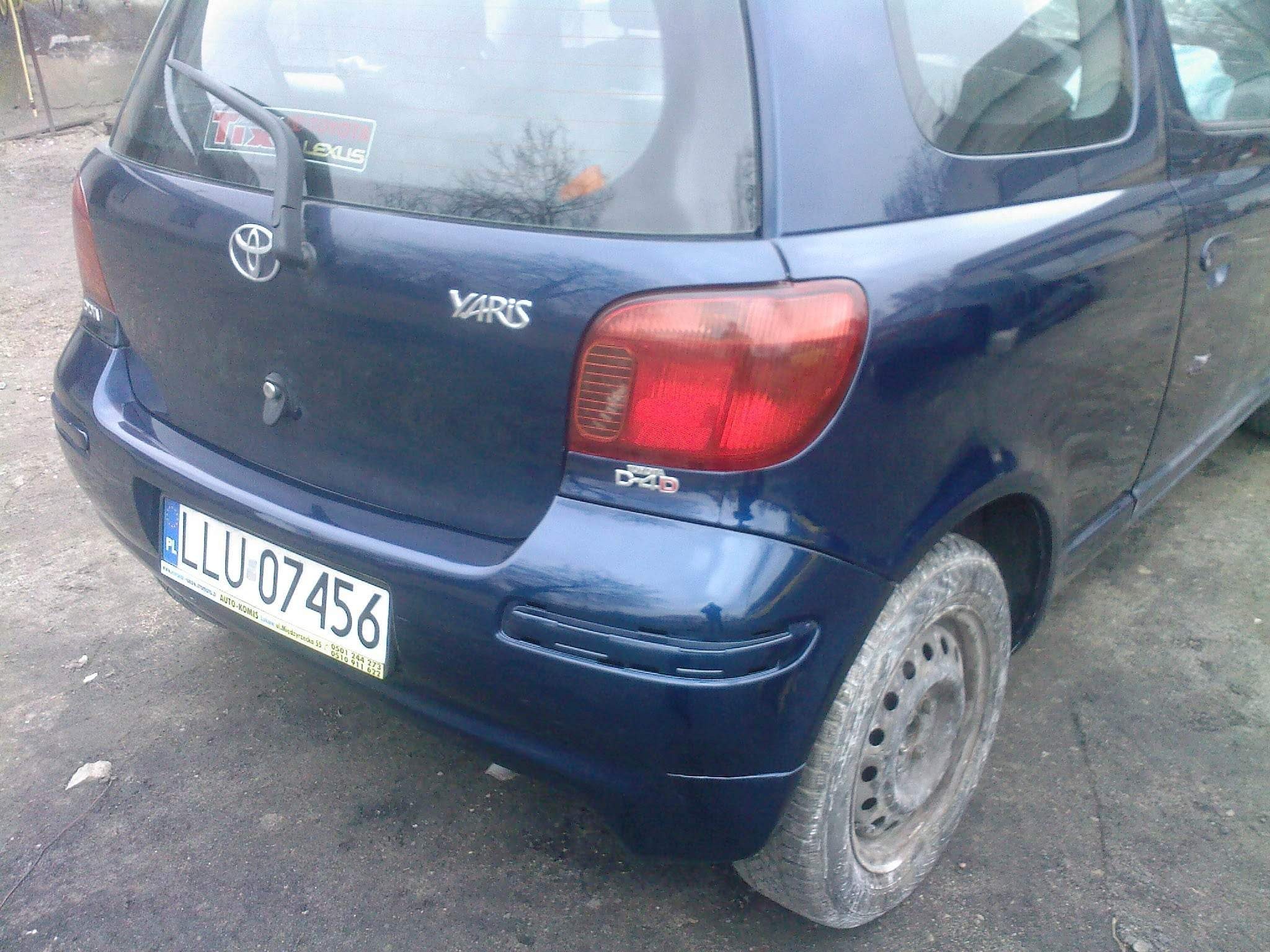 Tył granatowego samochodu Toyota Yaris z widoczną tablicą rejestracyjną, emblematem marki i naklejką na szybie, stojącego na brudnej nawierzchni.