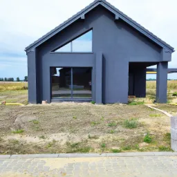 Nowoczesny, grafitowy dom jednorodzinny z dużymi oknami. Minimalistyczna bryła budynku na tle zieleni. Widoczne detale wykończenia i geometryczne kształty okien.