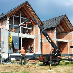 PWM TRADE MARCIN HAJZER - Montaż dużego okna za pomocą dźwigu przyssawkowego na placu budowy domu w stanie surowym otwartym, pracownicy w trakcie instalacji.