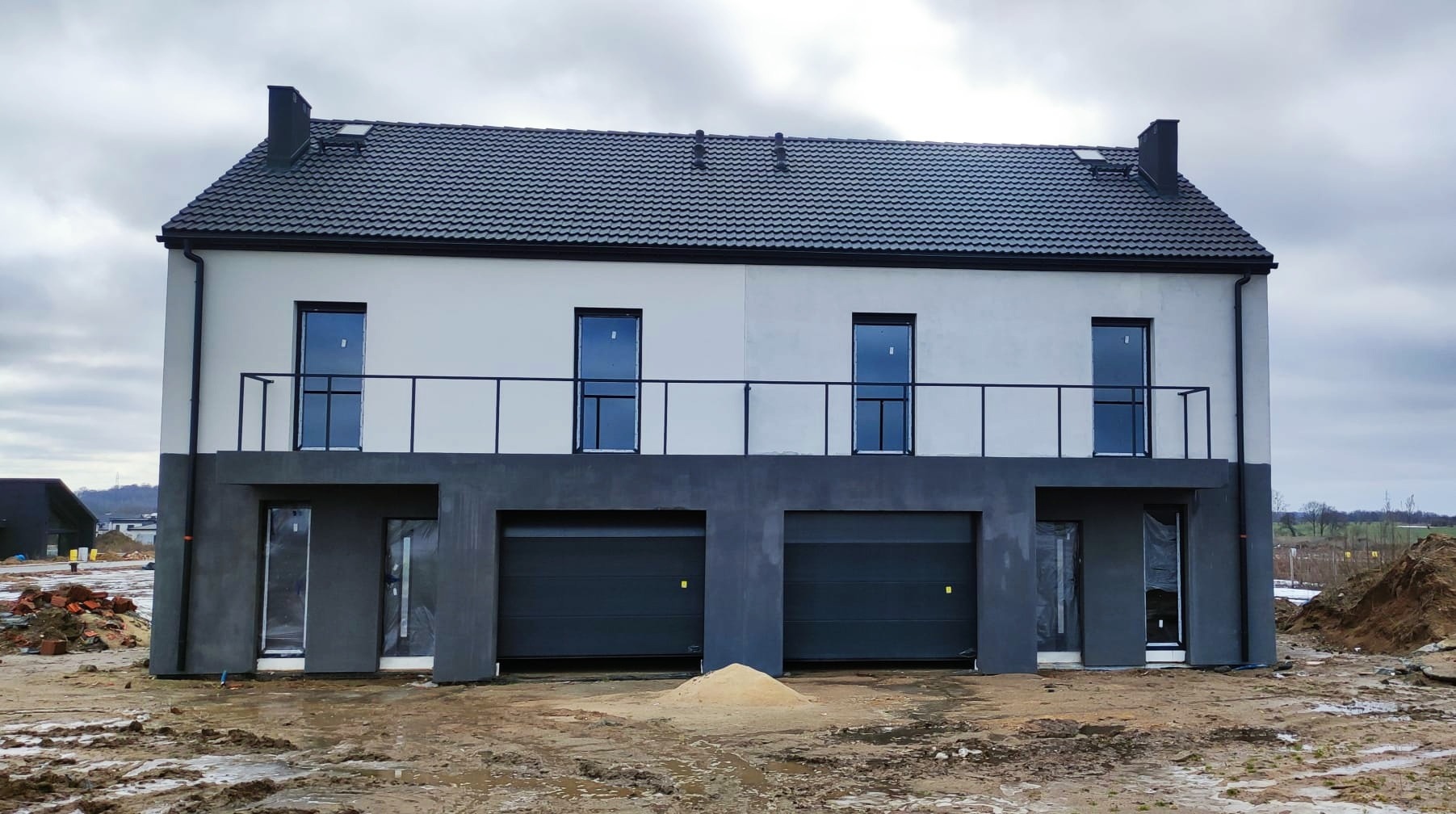 Nowoczesny, dwukondygnacyjny dom w trakcie wykończenia z szarymi bramami garażowymi i oknami na piętrze, widok z przodu na plac budowy.