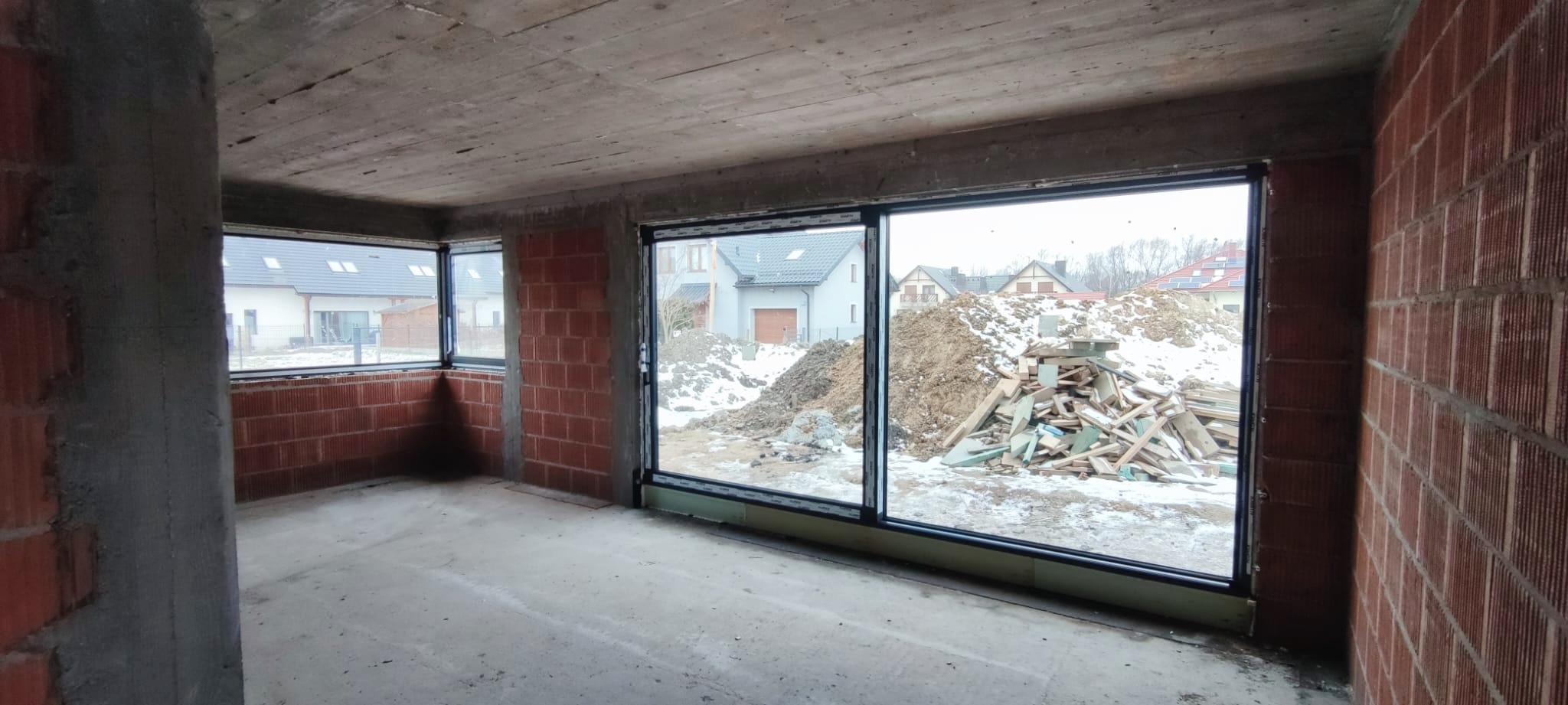 Wnętrze surowego budynku z zamontowanymi oknami PCV i dużym oknem przesuwnym. Widok na zasypany śniegiem plac budowy z hałdami ziemi i stertą drewna.