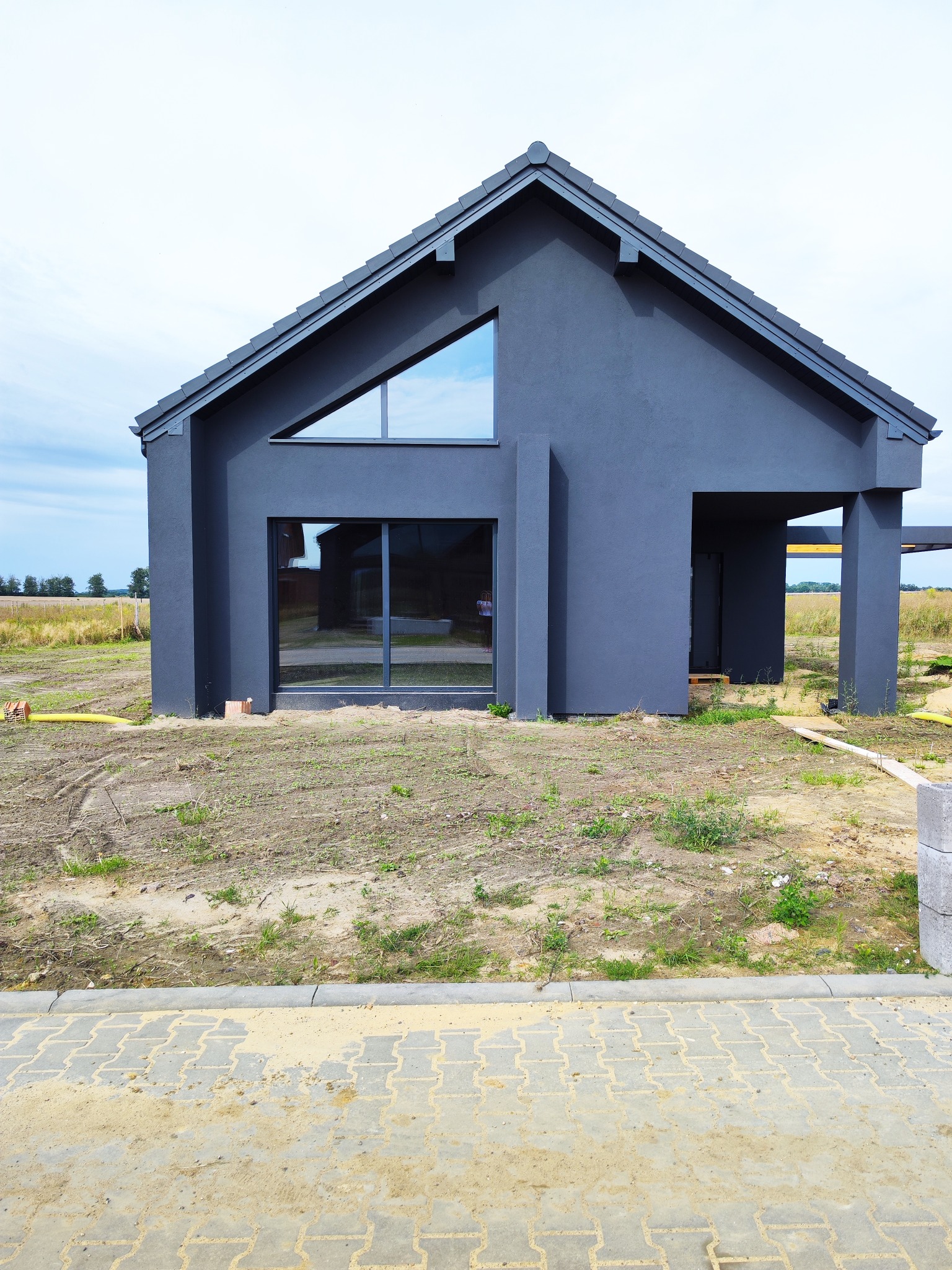 Nowoczesny, grafitowy dom jednorodzinny z dużymi oknami. Minimalistyczna bryła budynku na tle zieleni. Widoczne detale wykończenia i geometryczne kształty okien.