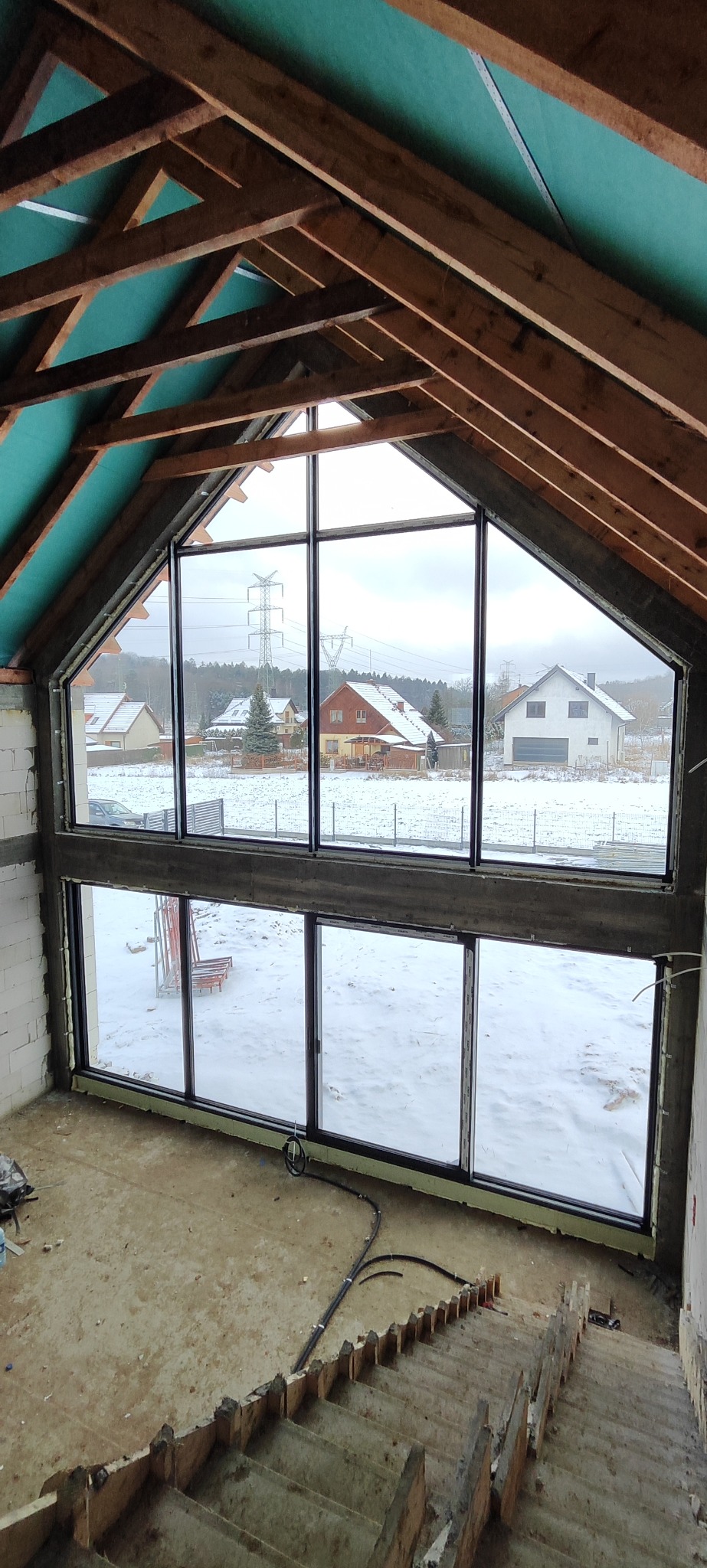 Duże okno aluminiowe w surowym domu z widokiem na zaśnieżony krajobraz. Widoczna konstrukcja dachu i schody. Ujęcie z góry, jasne światło dzienne.