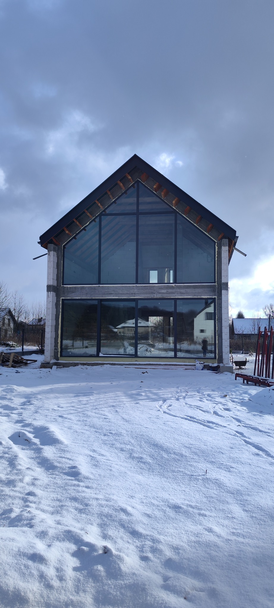 Surowy stan domu z dużymi oknami na dwóch kondygnacjach, w zimowej scenerii. Widoczne bloczki konstrukcyjne, drewniana konstrukcja dachu i refleksy w szybach.