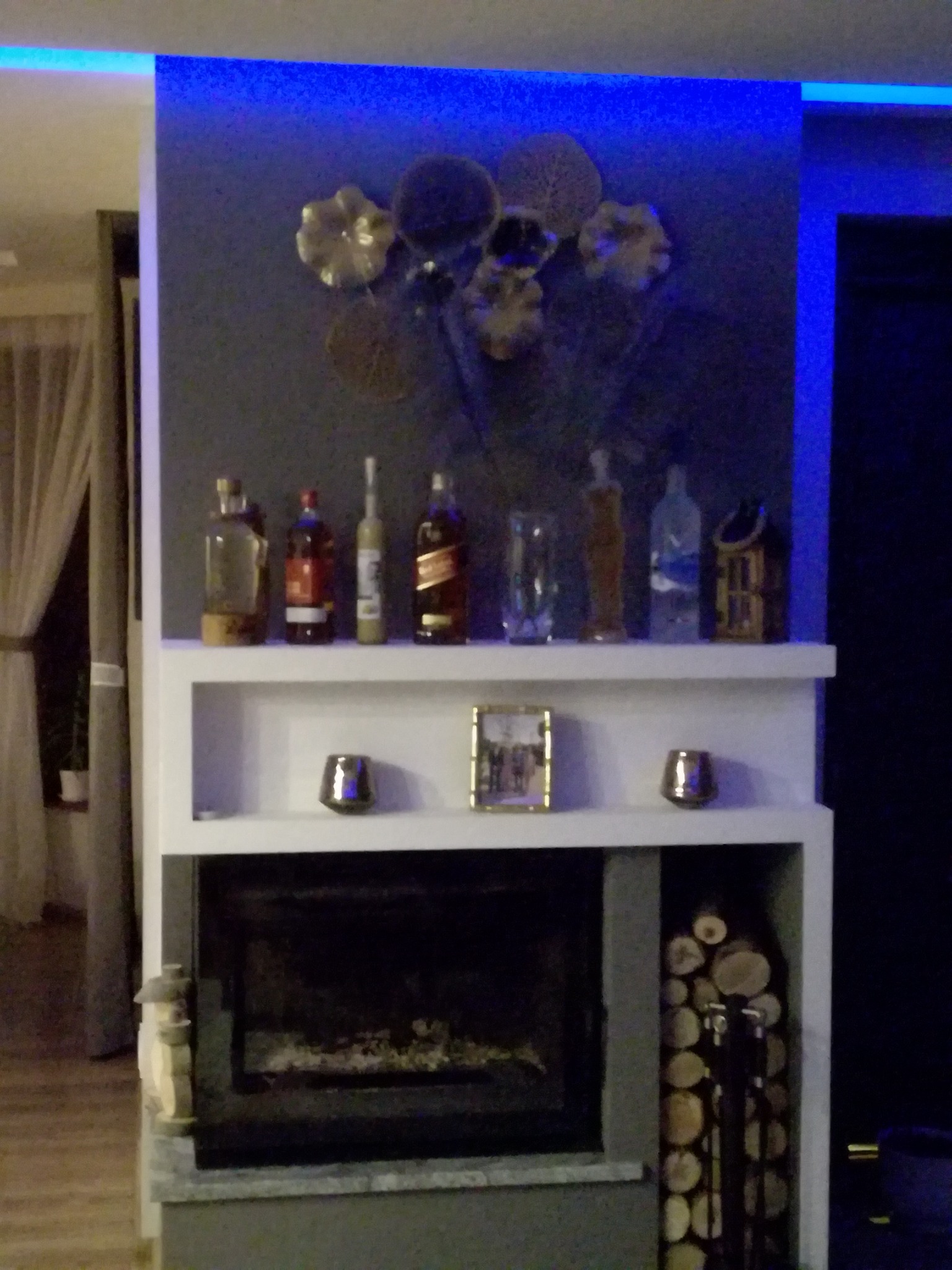 Nowoczesny kominek z półkami, podświetlony niebieskim światłem LED, na półkach dekoracyjne butelki i ozdoby, obok ułożone drewno kominkowe.