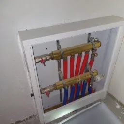 Rozdzielacz ogrzewania podłogowego w białej szafce, widoczne rury czerwone i niebieskie, zawory kulowe z czerwonymi uchwytami i termostat.