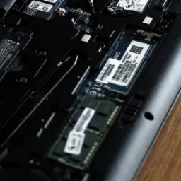 Wnętrze laptopa z widoczną pamięcią RAM i innymi komponentami na tle drewnianej powierzchni. Detaliczne ujęcie płyty głównej i zainstalowanych podzespołów.