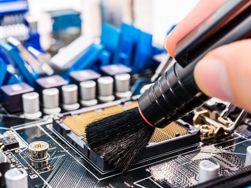 Czyszczenie gniazda procesora na płycie głównej komputera za pomocą pędzla antystatycznego, widoczne kondensatory i śruby mocujące.