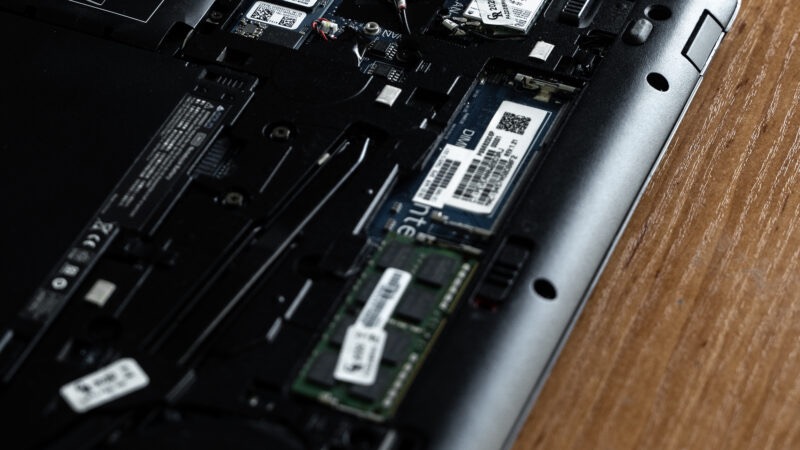 Wnętrze laptopa z widoczną pamięcią RAM i innymi komponentami na tle drewnianej powierzchni. Detaliczne ujęcie płyty głównej i zainstalowanych podzespołów.