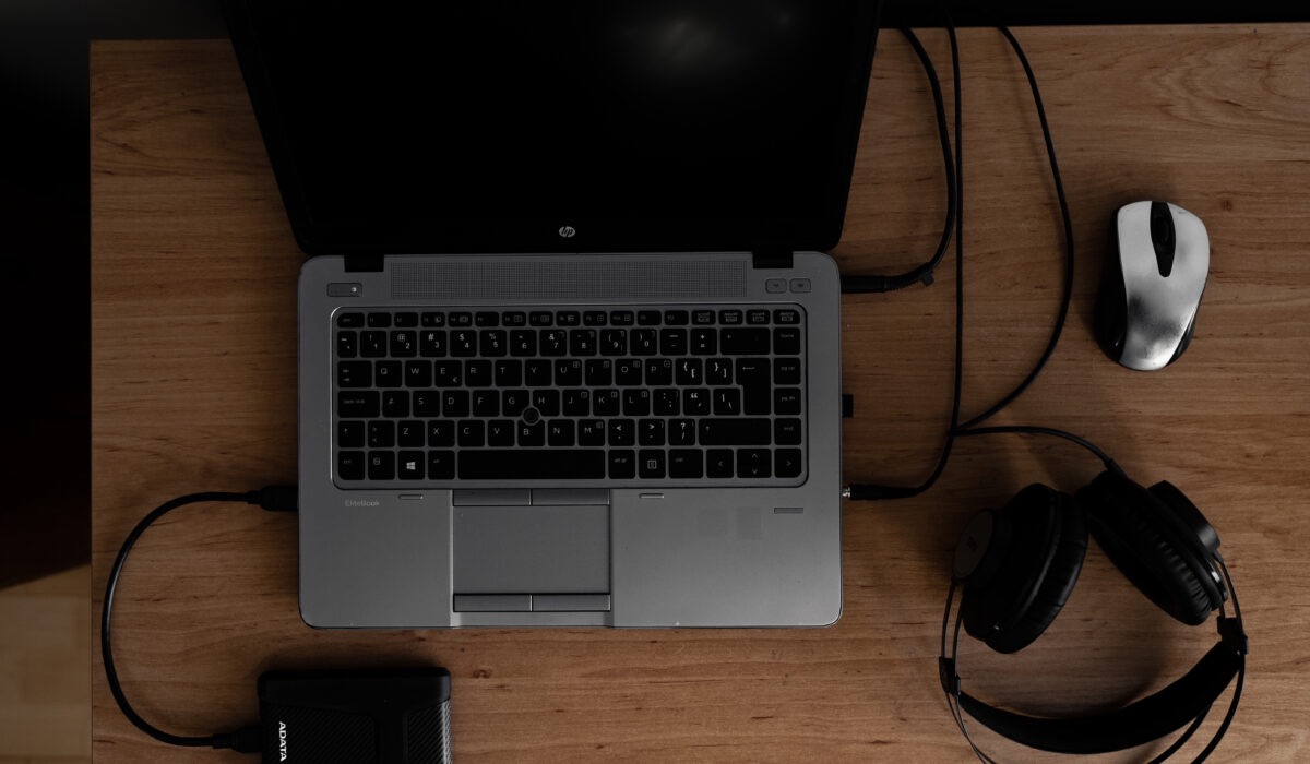 Widok z góry na otwarty laptop HP EliteBook, mysz, słuchawki i dysk zewnętrzny ADATA na drewnianym biurku. Kabel USB podłączony do komputera. Stonowane oświetlenie.