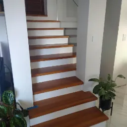 Nowoczesne schody drewniane z białymi podstopnicami, szklana balustrada na piętrze, rośliny doniczkowe po bokach, marmurowa podłoga.