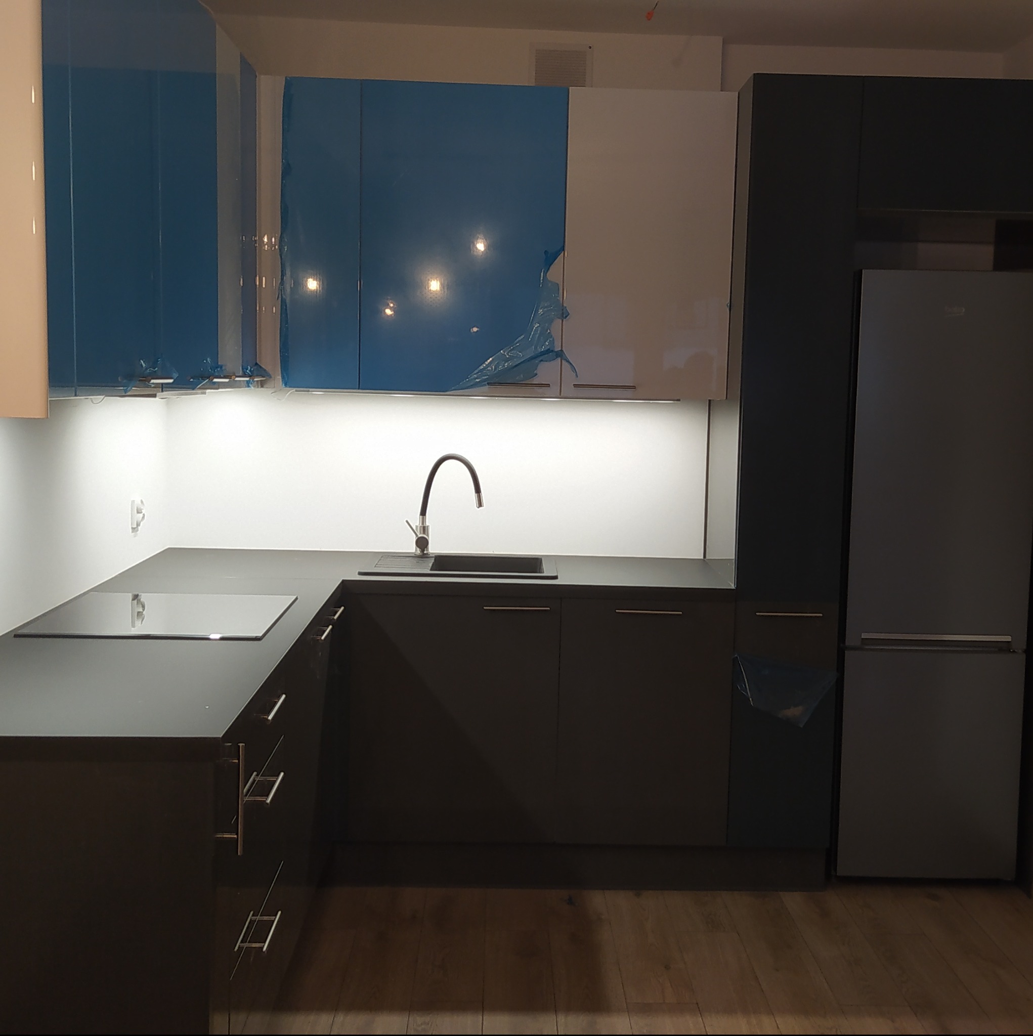 Nowoczesna kuchnia z szarymi szafkami i blatem, płyta indukcyjna i zlewozmywak z wysoką baterią, część szafek górnych zabezpieczona folią ochronną.