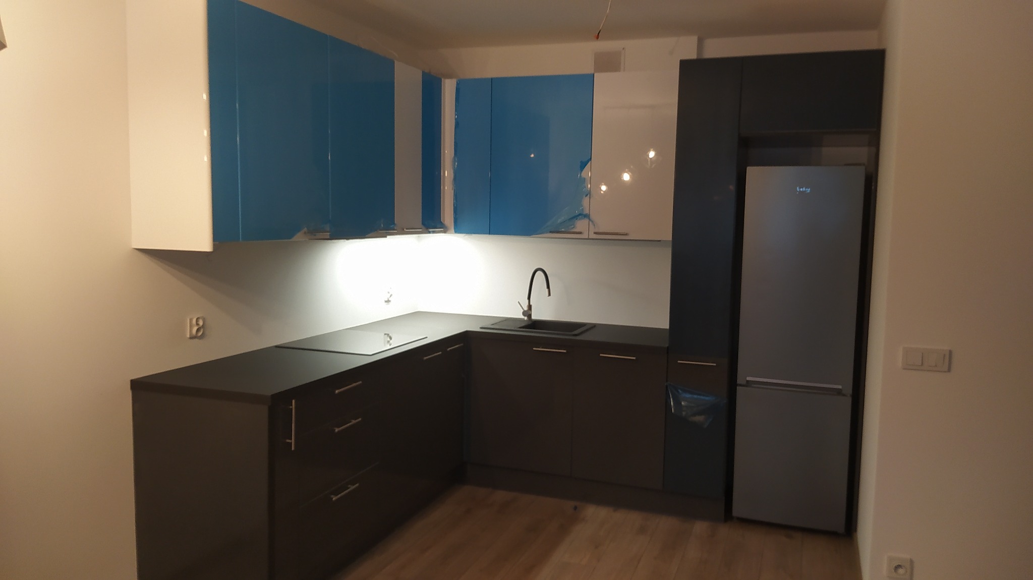 Nowoczesna kuchnia narożna z ciemnymi szafkami dolnymi, jasnoniebieskimi i białymi górnymi szafkami, z wbudowaną lodówką i płytą indukcyjną, częściowo jeszcze zabezpieczona folią ochronną.