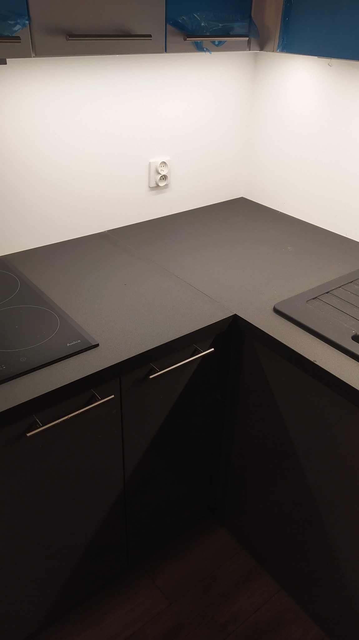 Czarny narożnik kuchenny z płytą indukcyjną Amica i zlewozmywakiem, widoczne szare szafki górne zabezpieczone folią ochronną, gniazdko elektryczne na ścianie.