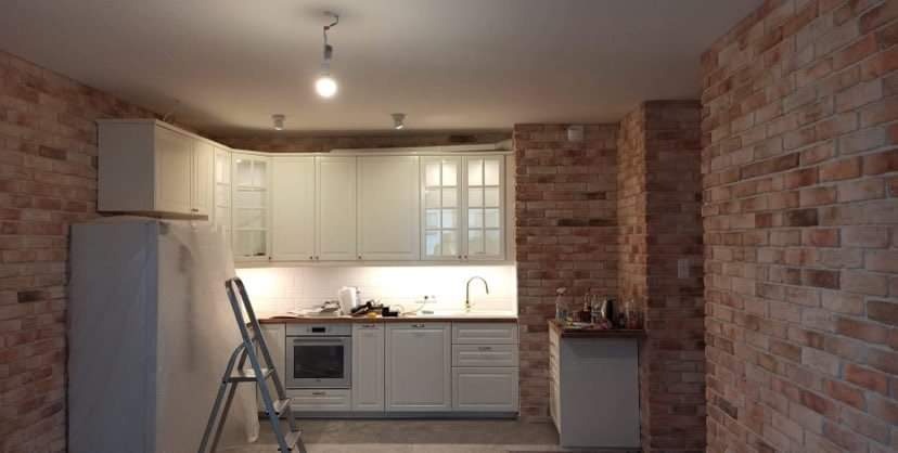 Wnętrze kuchni w trakcie montażu: białe szafki z przeszklonymi frontami, ceglana ściana, drabina oparta o lodówkę zabezpieczoną folią.