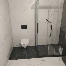 Nowoczesna łazienka z biało-czarnymi marmurkowymi płytkami, czarną armaturą i kabiną prysznicową typu walk-in z czarnymi profilami, wisząca toaleta z czarnym przyciskiem spłukującym.